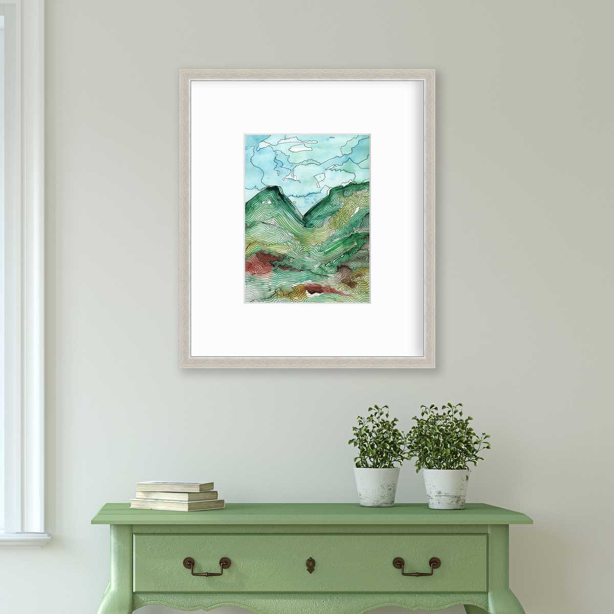 A blue, green and black abstract landscape print in a silver frame with a mat hangs on a white wall above a green console table with a stack of books and two potted plants.