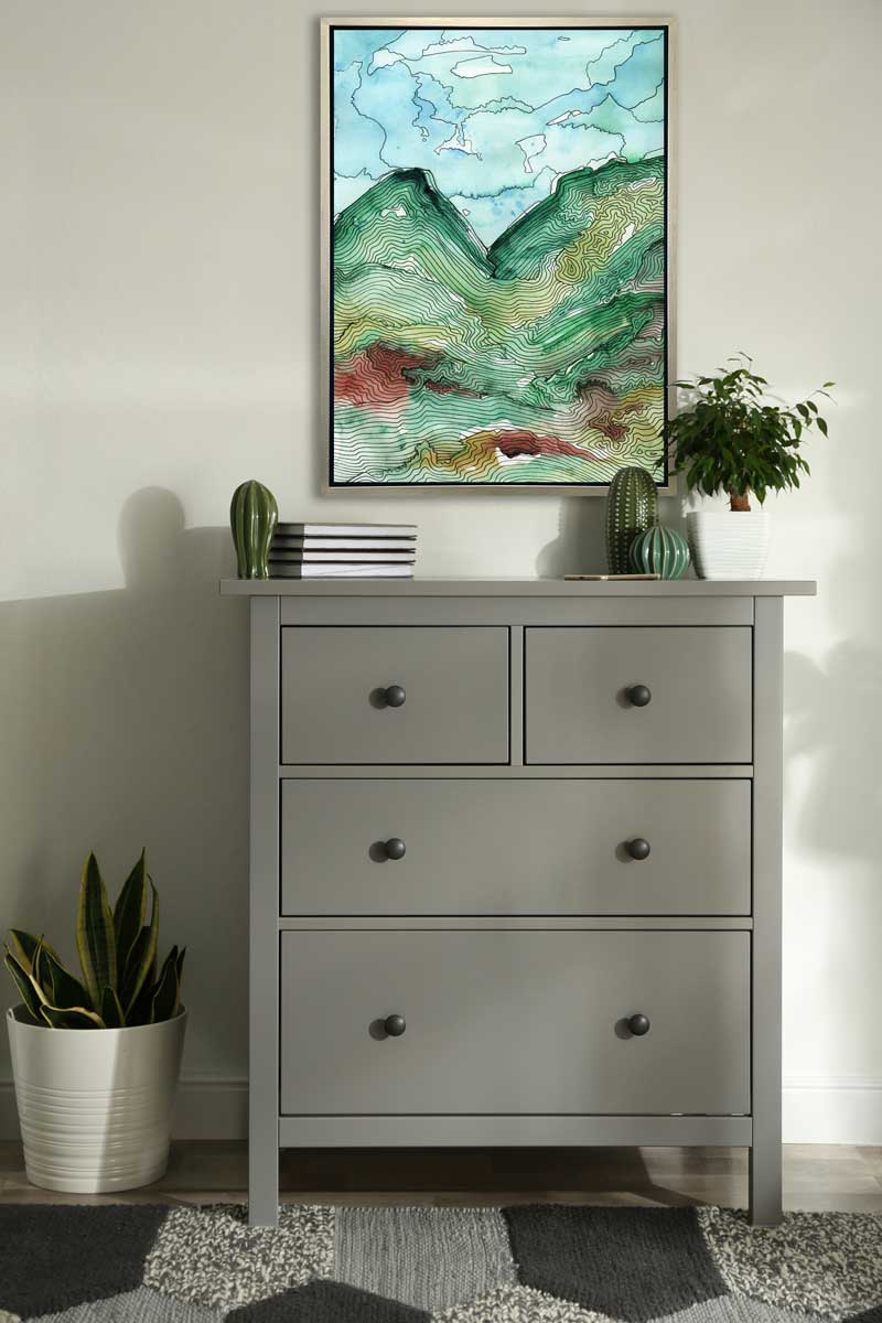 A blue, green and black abstract landscape print in a silver floater frame hangs on a white wall above a light grey dresser, small cacti decorations, a stack of four books and a small potted plant.