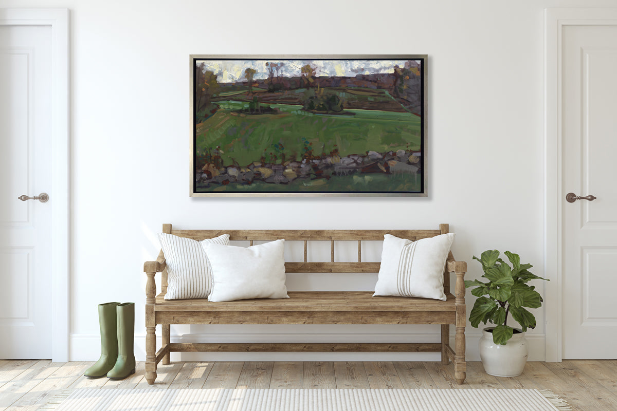 A green, brown and white abstract landscape print in a silver floater frame hangs on white wall above a wood bench with white pillows, a potted floor plant and a pair of green rubber boots.