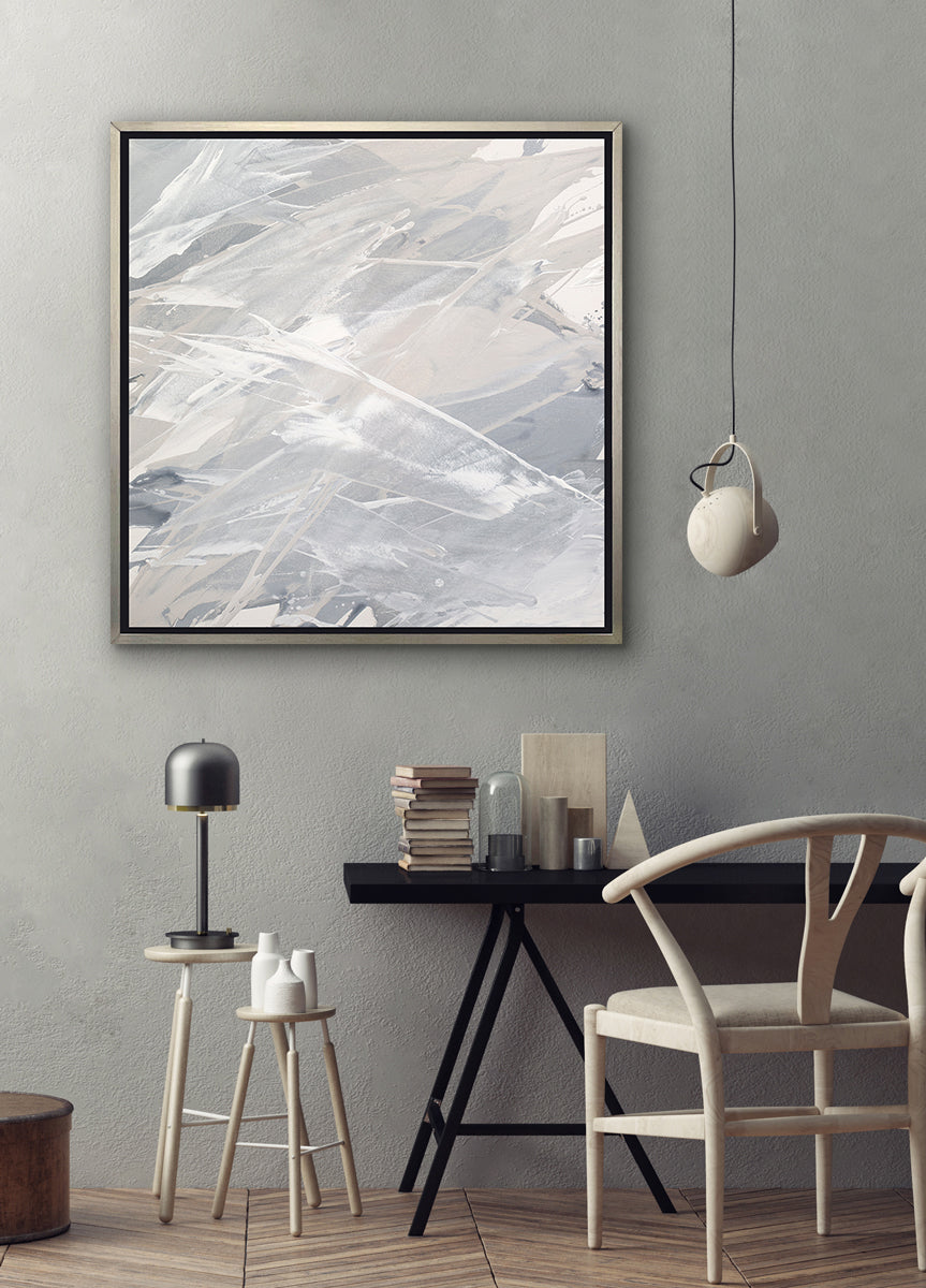 A grey, beige and white abstract print in a silver floater frame hangs on a grey wall above a black desk with a stack of books, wood decorations, a light wood chair and wood stools with decorative accessories.