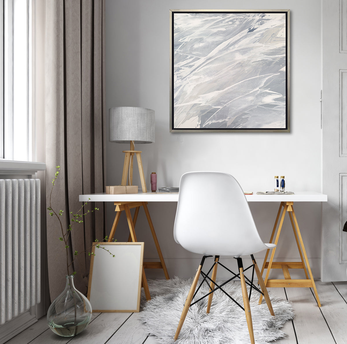 A grey, beige and white abstract print in a silver floater frame hangs on a white wall above a white desk, with a wood tripod desk lamp, sewing thread, a white desk chair and a glass vase on the floor with a greenery stem.