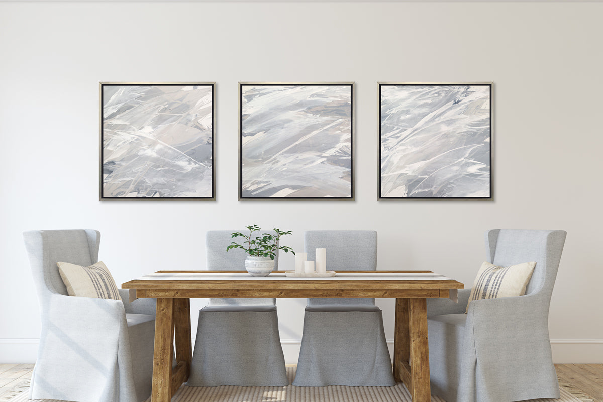 Three grey, beige and white abstract prints in silver floater frames hang on a white wall above a wood table with a potted plant and four high back grey chairs
