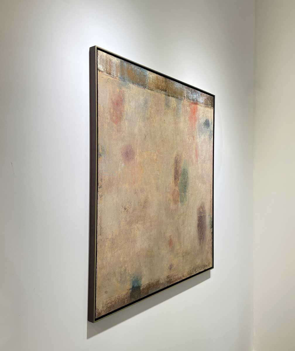 A left side-angle view of a beige painting with green, blue and purple accents in a warm gold floater frame hangs on a white wall.