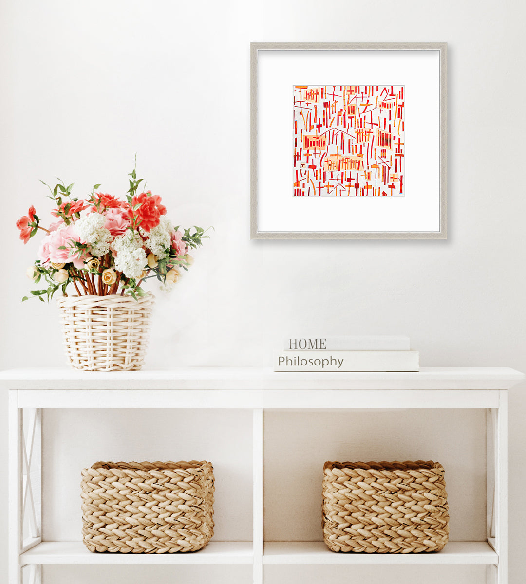 A red, orange and white abstract print in a silver frame with a mat hangs on a white wall above a white console table with a stack of books, a woven potted floral arrangement and two woven baskets.
