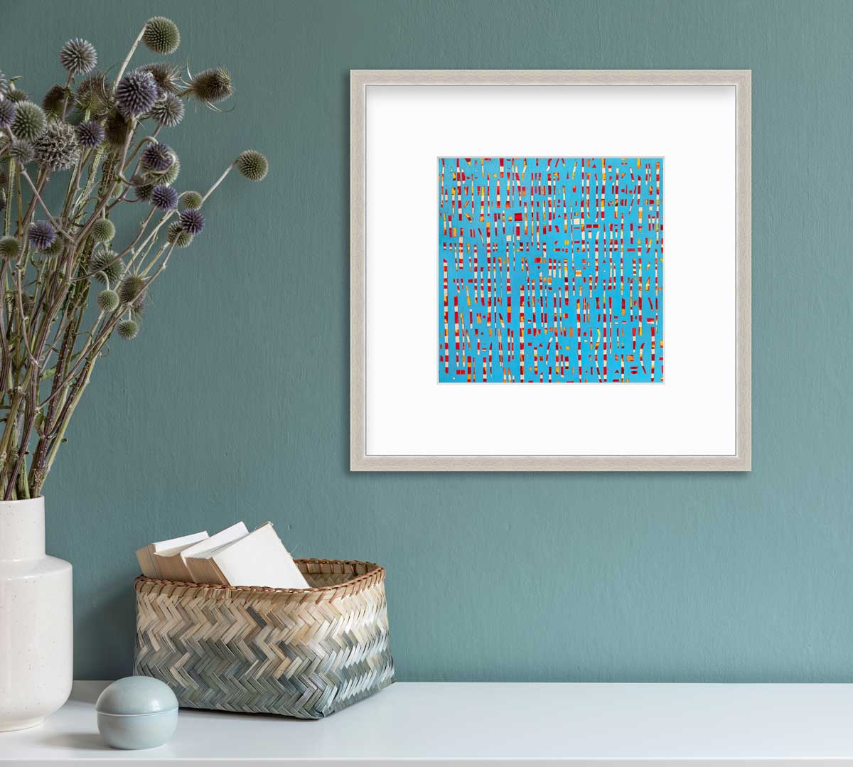 A blue, red, yellow and white abstract print in a silver frame with a mat hangs on a white wall above a white surface with a woven beige and blue basket with card stock, a small rounded light blue container and a white vase with green and purple pom pom stems.