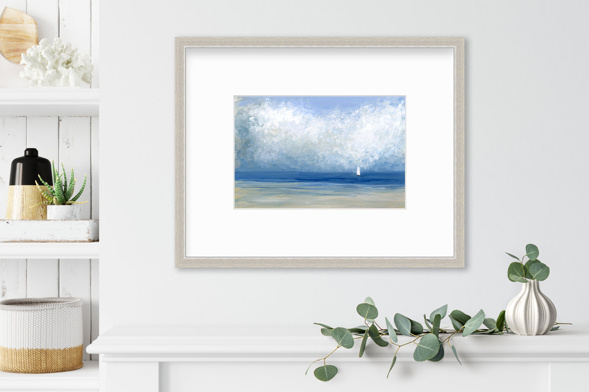 A blue, white and beige abstract seascape print in a silver frame with a mat hangs on a white wall above a white shelf with a eucalyptus filled white vase and next to a built in bookshelf with decorative accessories.