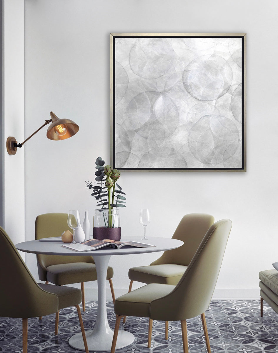 A grey and white abstract geometric print in a silver floater frame hangs on a white wall above a white pedestal table with a vase of succulents, some glassware, and an open magazine. Four olive green chairs surround the table.
