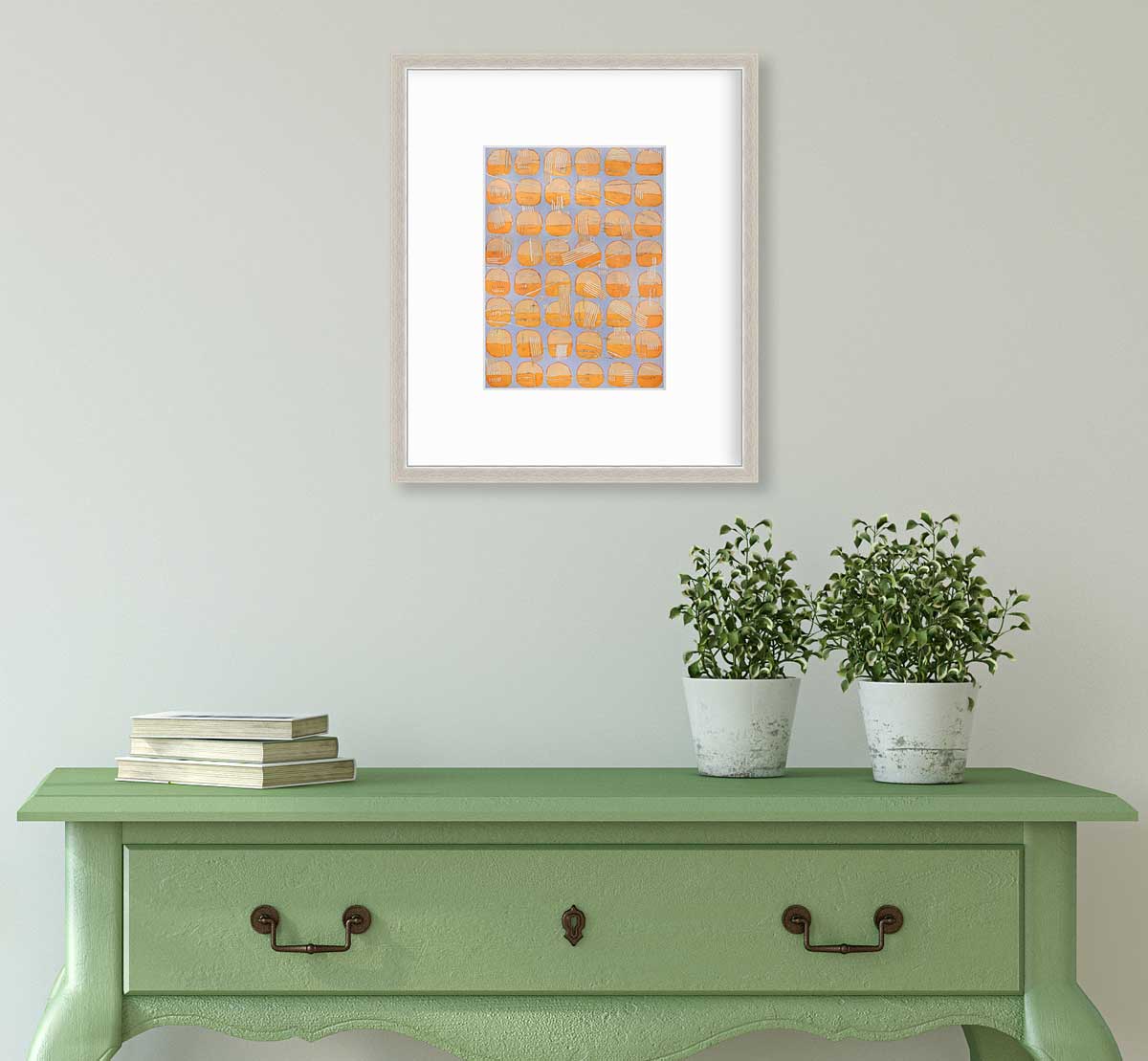 A orange and light purple abstract circle print in a silver frame with a mat hangs on a white wall above a green console table with a stack of books and two potted plants.
