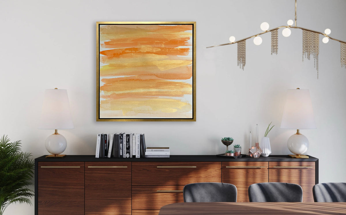 A orange, gold and white abstract print in a gold floater frame hangs on a white wall above a wood sideboard with a stack of books, a set of matching white table lamps and succulent arrangements.