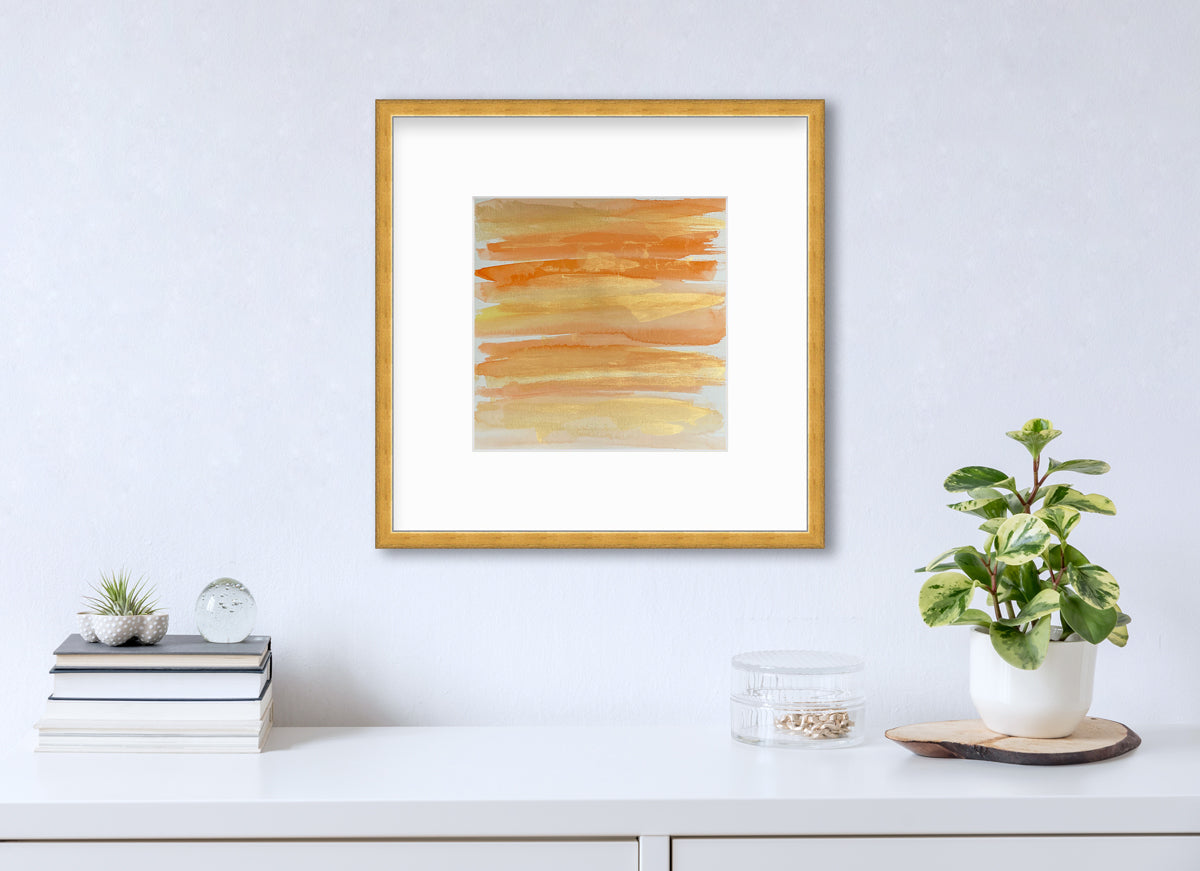 A orange, gold and white abstract print in a gold frame with a mat hangs on a white wall above a white surface with a stack of books, a round clear container with gold accessories and a potted plant.