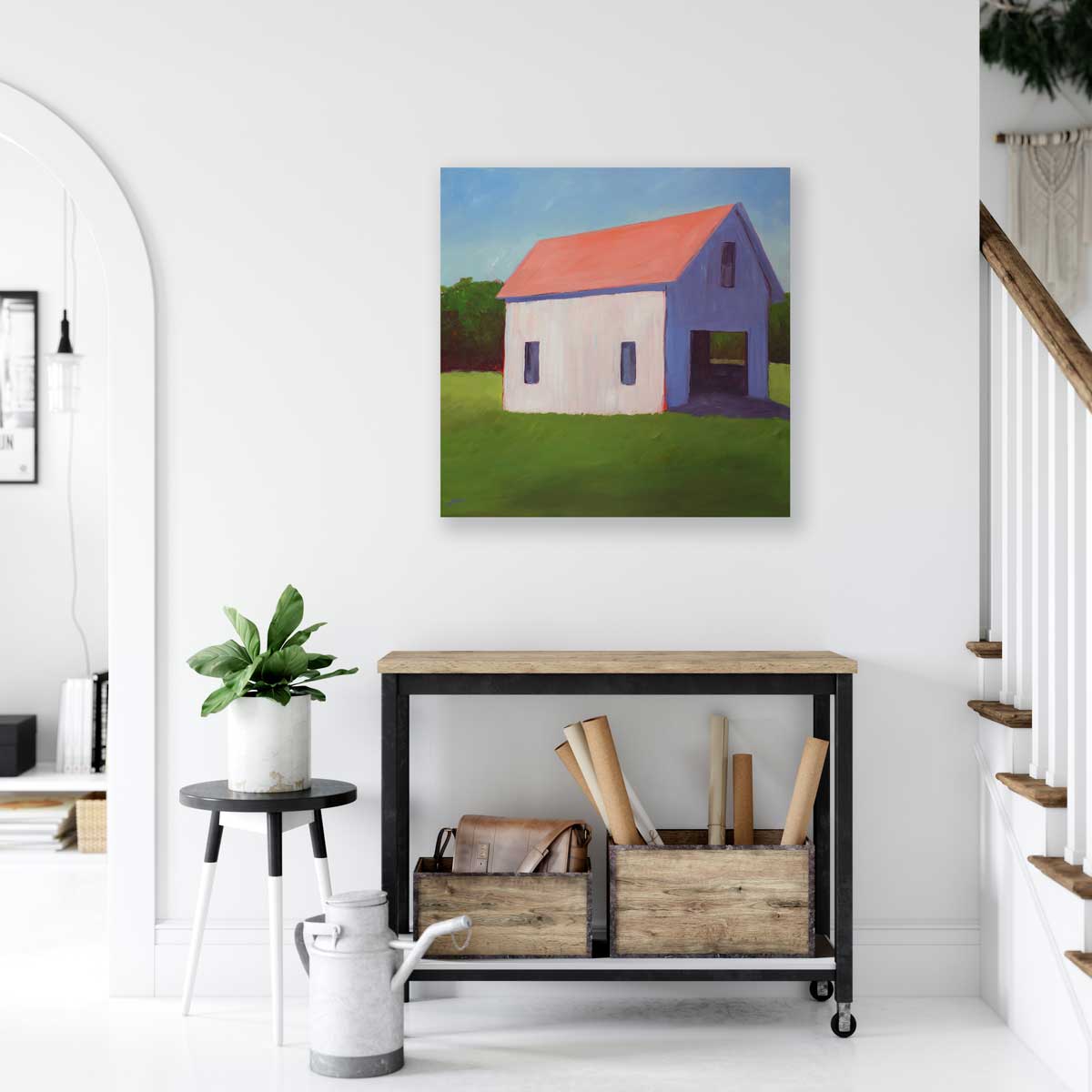 A painting of a white barn with a salmon roof in a green field with a pale blue sky background hanging on a white wall above a black rolling console with a natural wood top and baskets underneath to the left of a staircase. 