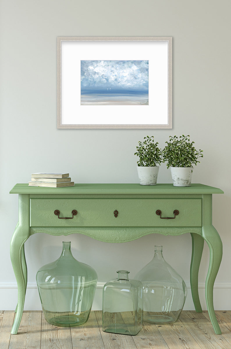 A white, blue and beige abstract seascape print in a silver frame with a mat hangs on a white wall above a green console table with a stack of books, two potted plants and three large green glass vases.