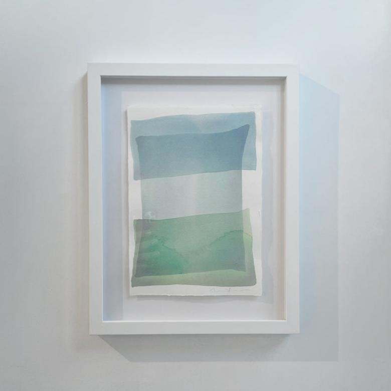 Tonal blues and green shapes overlap in this painting by Nealy Hauschildt in a white frame floating above a white matte hangs on a white wall.