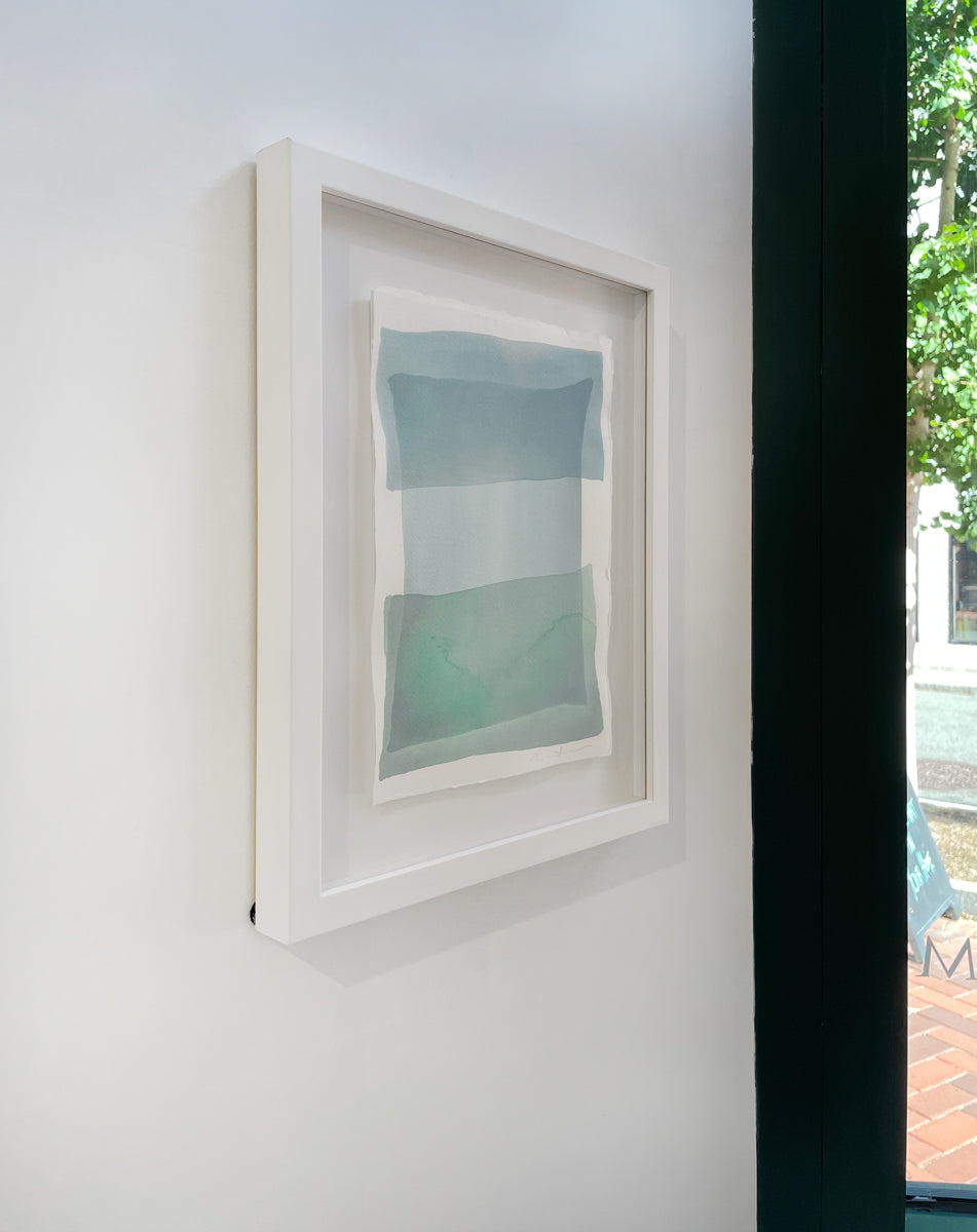 A left side-angle view of tonal blues and green shapes overlap in this painting by Nealy Hauschildt in a white frame floating above a white matte hangs on a white wall.