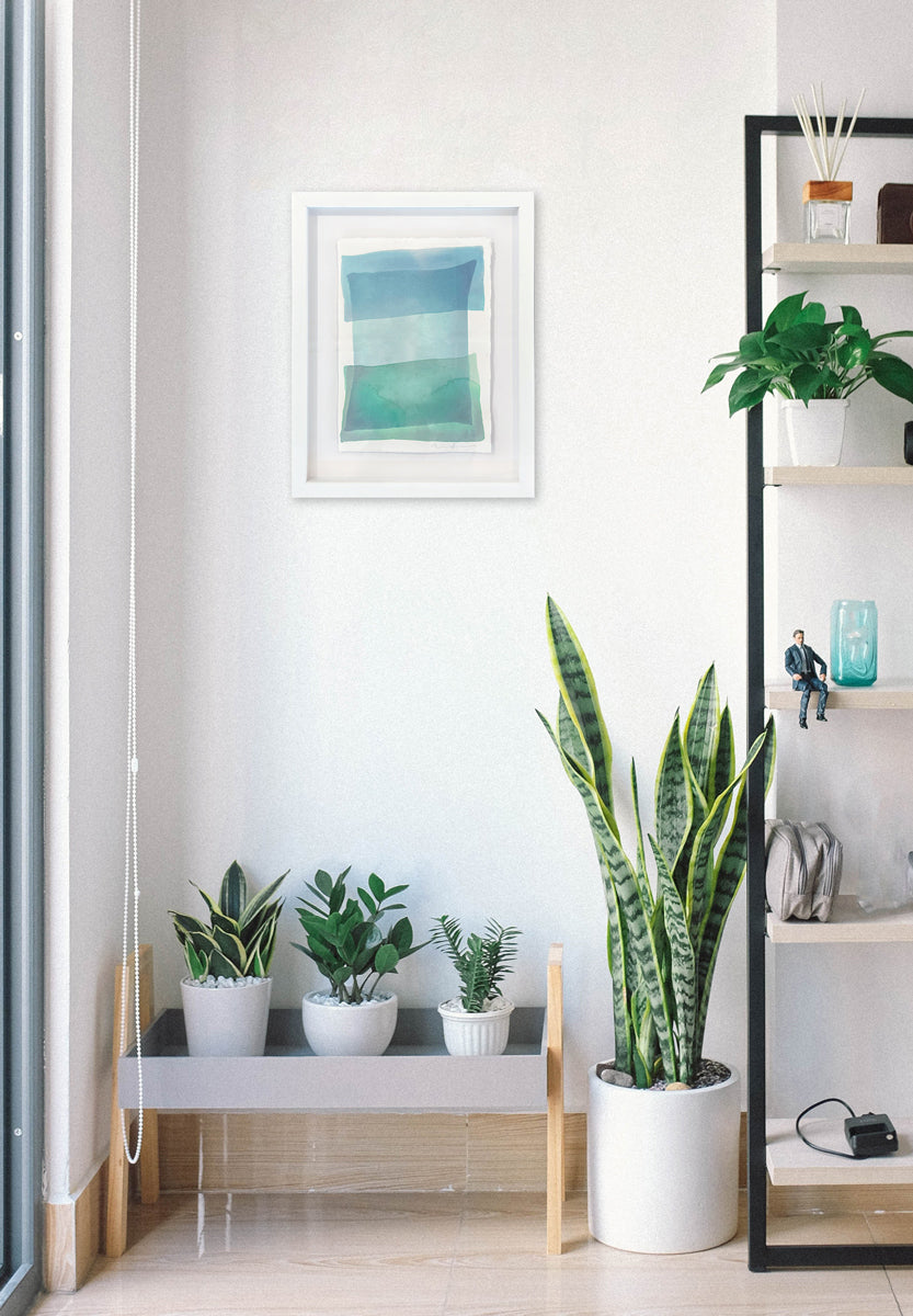 Tonal blues and green shapes overlap in this painting by Nealy Hauschildt in a white frame floating above a white matte hangs on a white wall above a small bench with potted plants, a medium potted plant on the floor and a tall ladder bookshelf with decorative accessories.