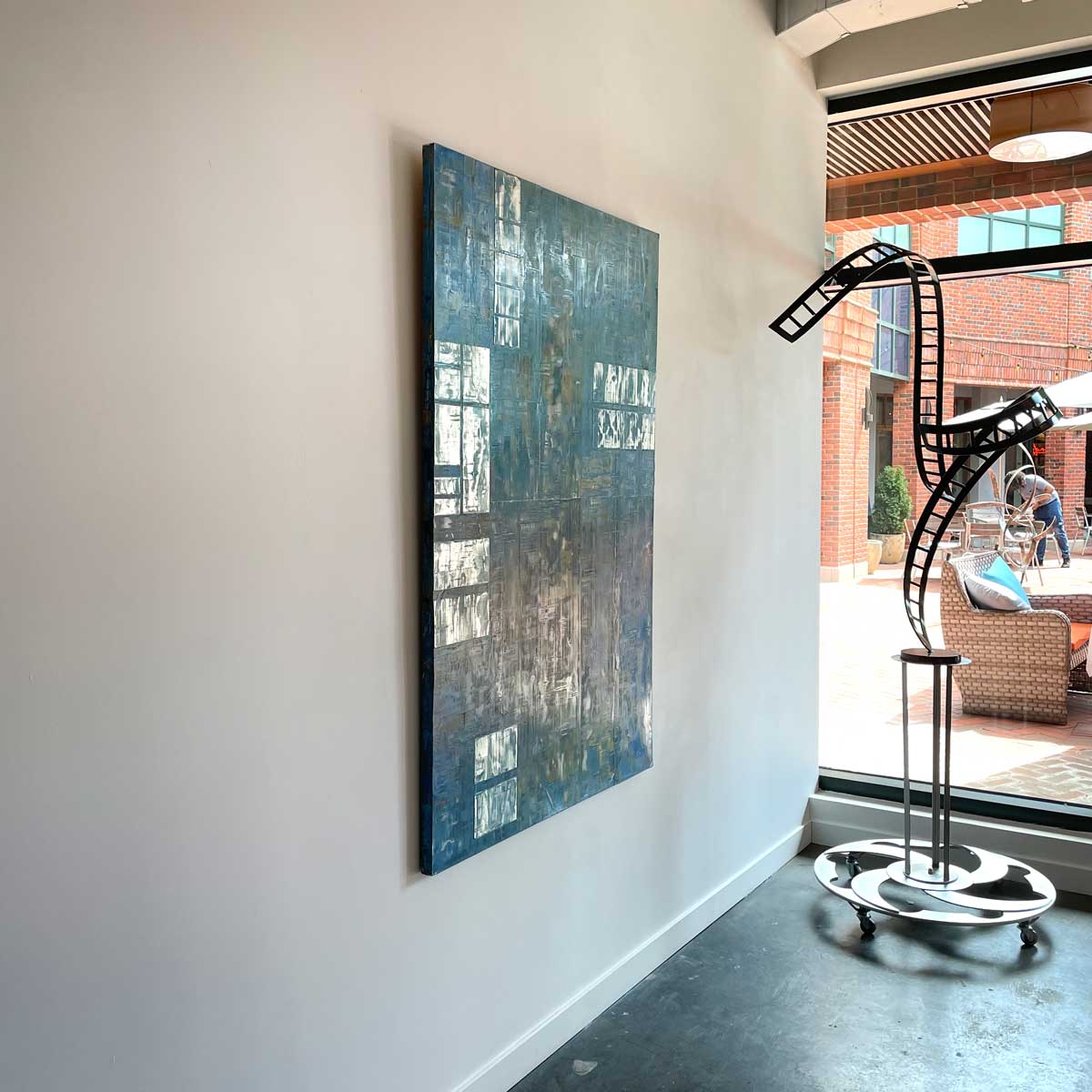 Mixed media abstract painting with small panels of aluminum and blue wash by Ned Martin hangs on a white wall with a view from the left. 