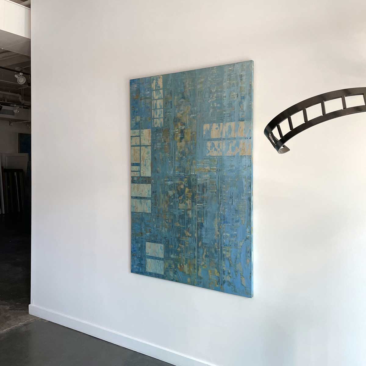 Mixed media abstract painting with small panels of aluminum and blue wash by Ned Martin hangs on a white wall with a view from the right. 