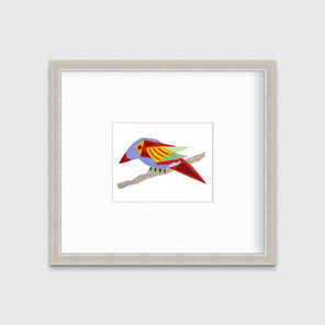 A multicolored bird on a branch collage print in a silver frame with a mat hangs on a white wall.