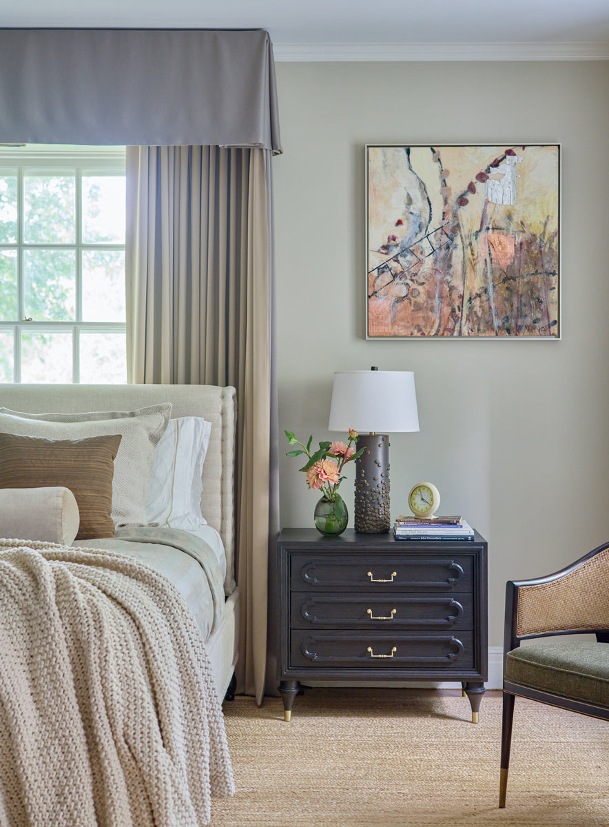 An earth-toned abstract encaustic painting by Linda Bigness hangs on a beige wall in a master bedroom. The painting hangs to the right of the king size bed with neutral bedding and above an espresso night stand. On top of the night stand is a stack of books, a floral arrangement in a vase, and a brown textured cylindrical lamp.  