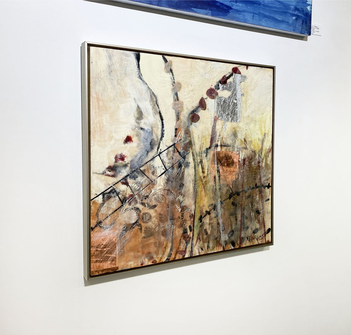 A left-hand side-angle view of a nature-inspired abstract encaustic painting, framed in a thin, warm silver floater frame hanging on a white wall. 