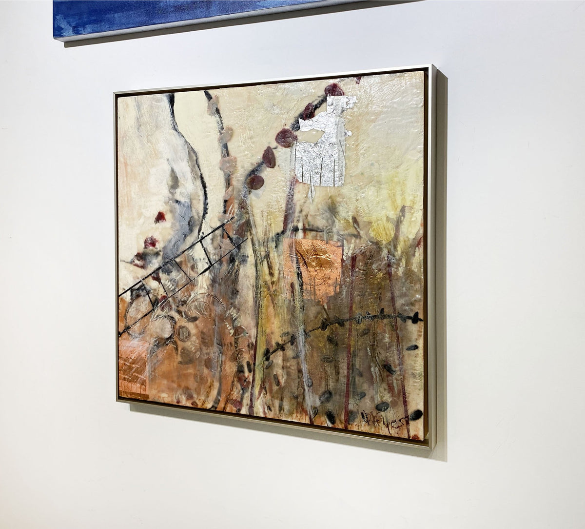 A right-hand side-angle view of a nature-inspired abstract encaustic painting, framed in a thin, warm silver floater frame hanging on a white wall.