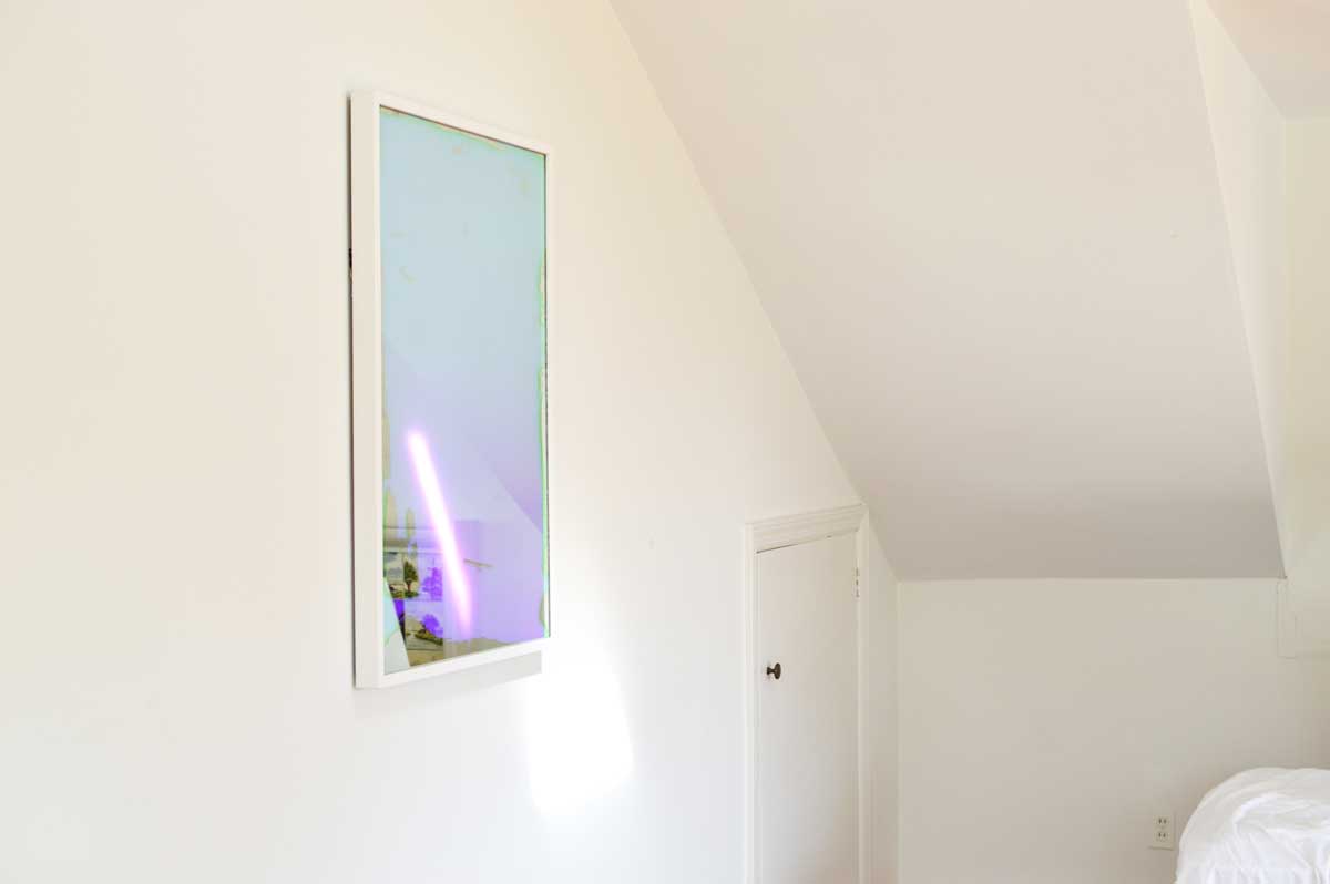 A multicolored fine art mirror hangs on a white wall near corner with a steep ceiling.