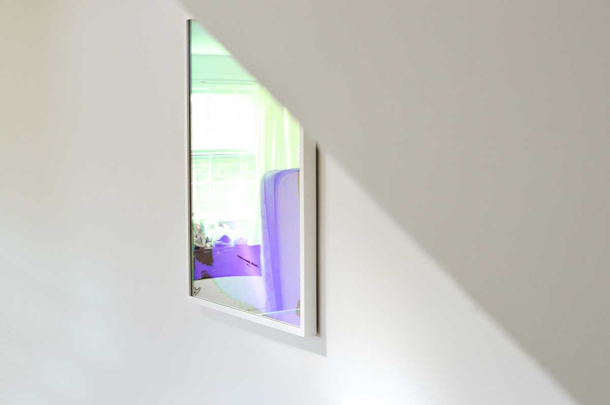 A fine art mirror by artist Alina B hangs on a white wall, partly obscured from view by a low ceiling. 