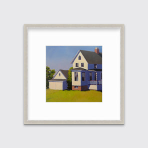 A blue, white and green contemporary house print in a silver frame with a mat hangs on a white wall.