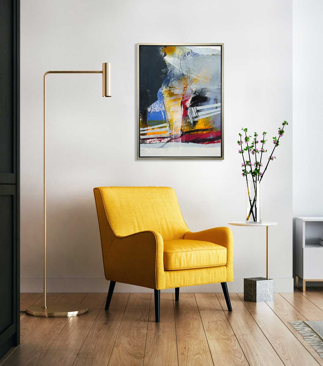 A white, yellow, red and grey abstract print in a silver floater frame hangs on a white wall above a yellow chair, a white marble side table with a clear vase with floral stems and a tall floor lamp.