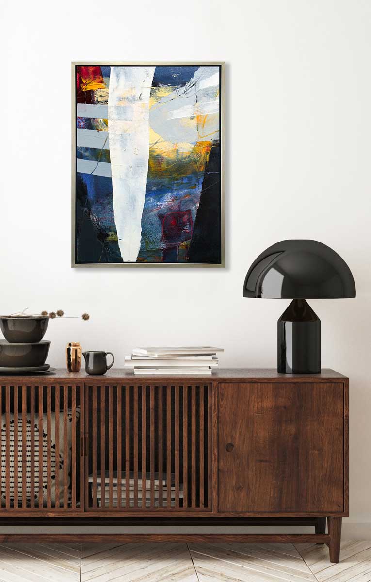 A white, yellow, red and grey abstract print in a silver floater frame hangs on a white wall above a stained wood sideboard with a black modern table lamp, a stack of books and a stack of dishes.