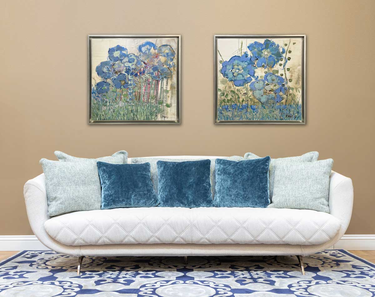 Two abstract, encaustic floral paintings by Linda Bigness hang on a brown wall in a warm silver floater frame above a cream sofa with velvet blue pillows.