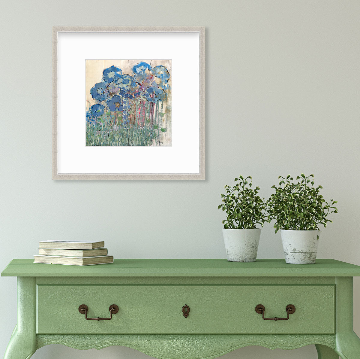 A blue and green abstract floral print in a silver frame with a mat hangs on a white wall above a green console table with two potted plants and a stack of books.