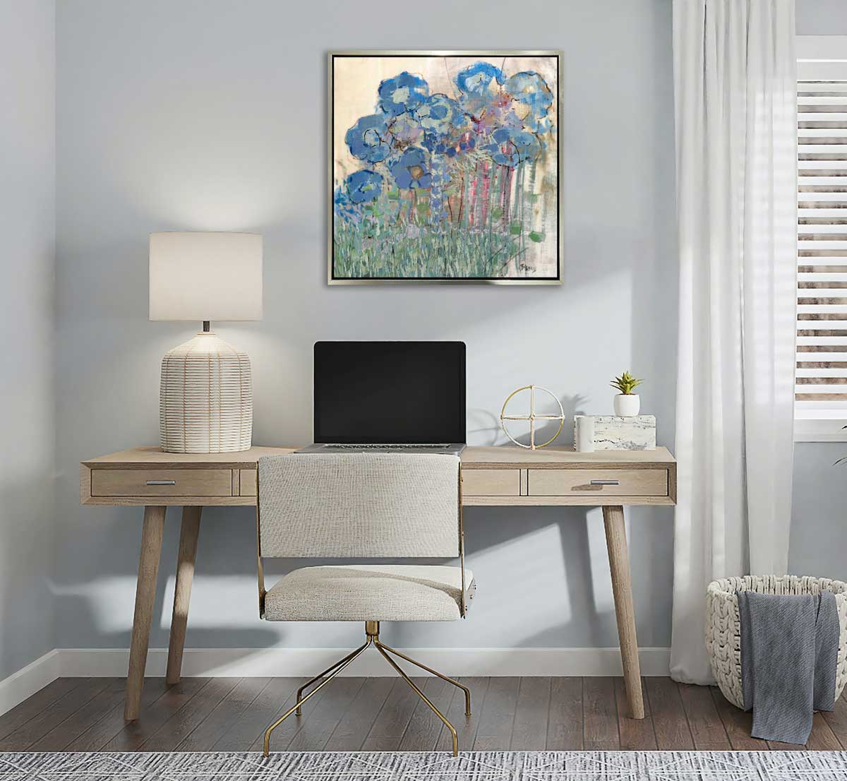 A blue and green abstract floral print in a silver floater frame hangs on a white wall above a wooden desk with a table lamp, a laptop computer and small accessories.