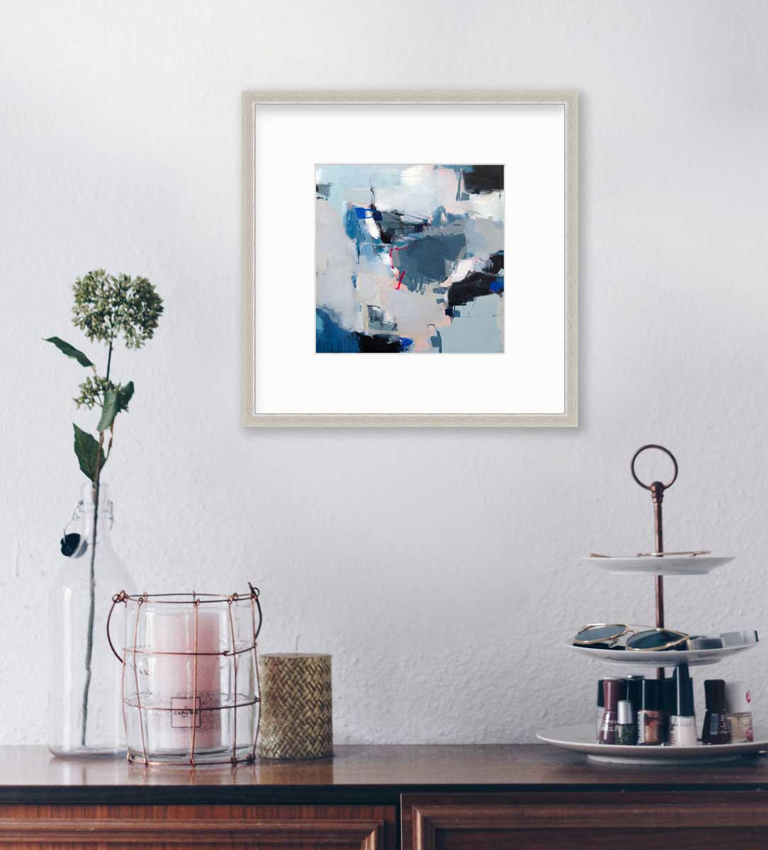 A blue abstract print by Kelly Rossetti in a silver frame with a mat hangs on a white wall above a dark wood countertop with a three-tiered caddy with nail polish, two candleholders and a clear bottle with a floral stem.