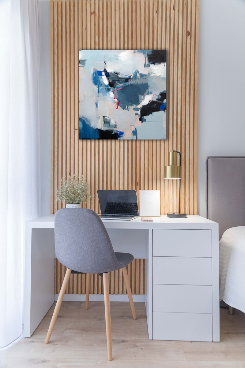 A blue, white and black abstract painting hangs above a small white desk with drawers to the right and a grey desk chair.
