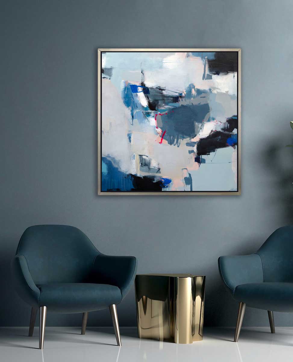 A blue, white and black abstract print in a silver floater frame hangs on a muted teal wall above two dark teal chairs and a gold decorative table.