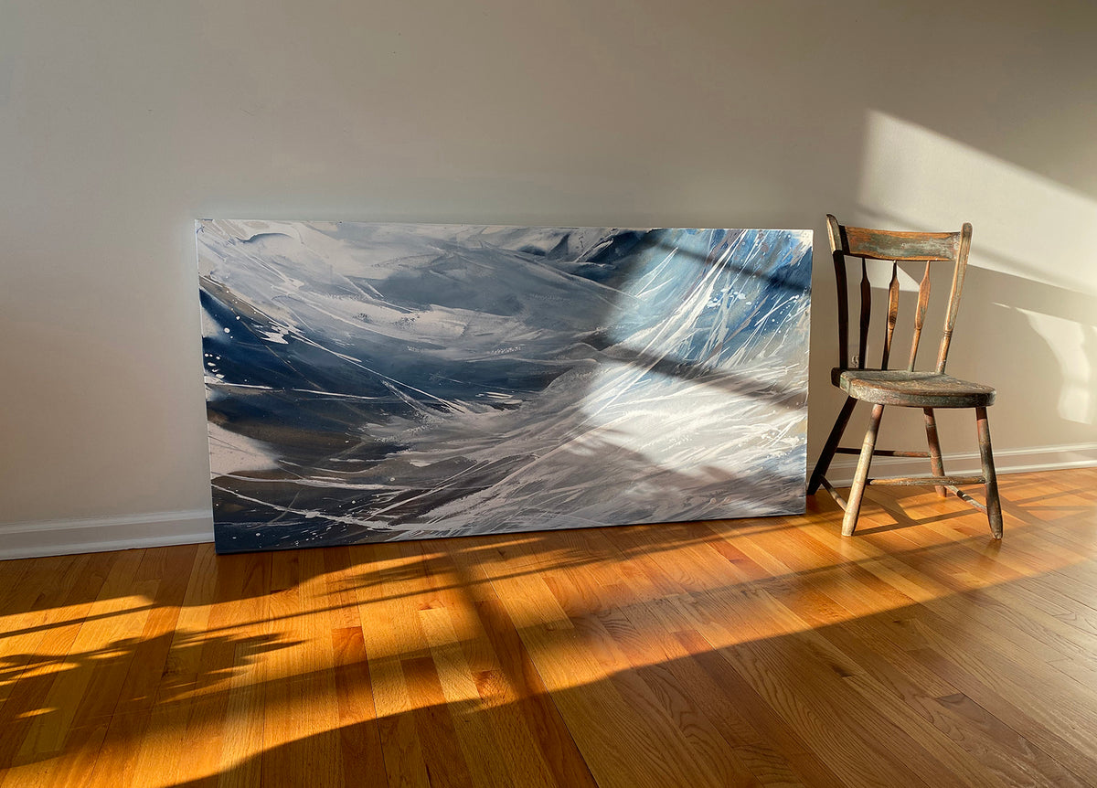 A white, blue, and grey abstract painting by Teodora Guererra sits on a hardwood floor with the sun cascading in. A wooden chair sits to the right of the painting.