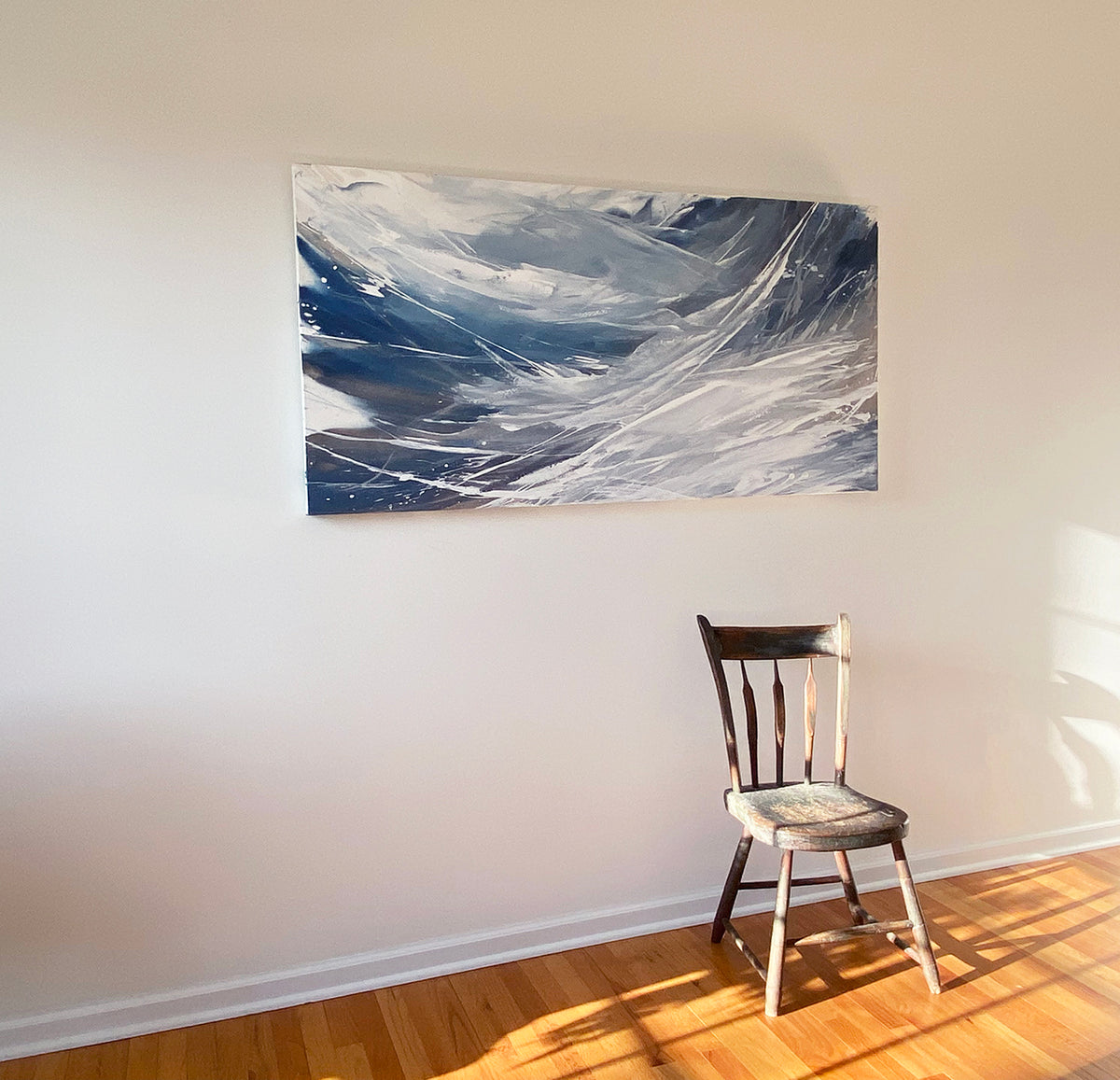 A white, blue, and grey abstract painting by Teodora Guererra hangs on a white wall with the sun shining in.  A wooden chair sits below the painting.tin.