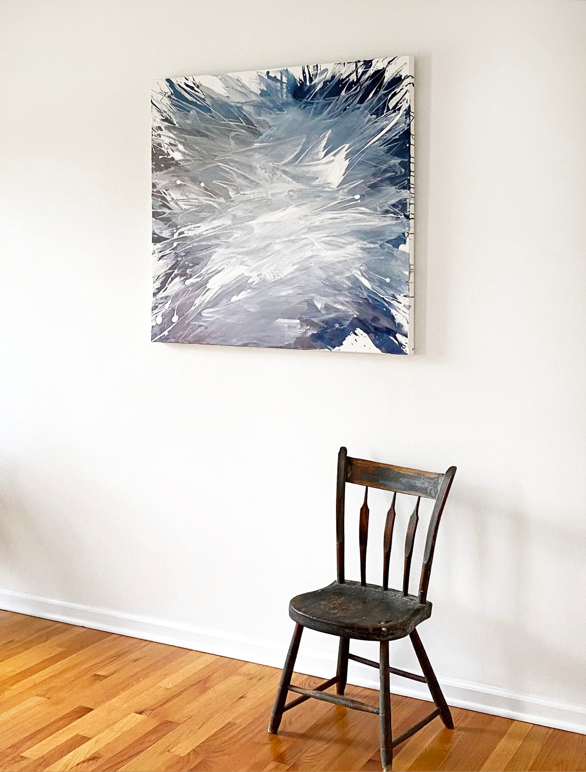 A white, blue, and lavender abstract painting by Teodora Guererra hangs on a white wall with a wooden chair sitting below. 