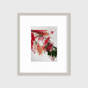 A red, green and white abstract print in a silver frame with a mat hangs on a white wall.