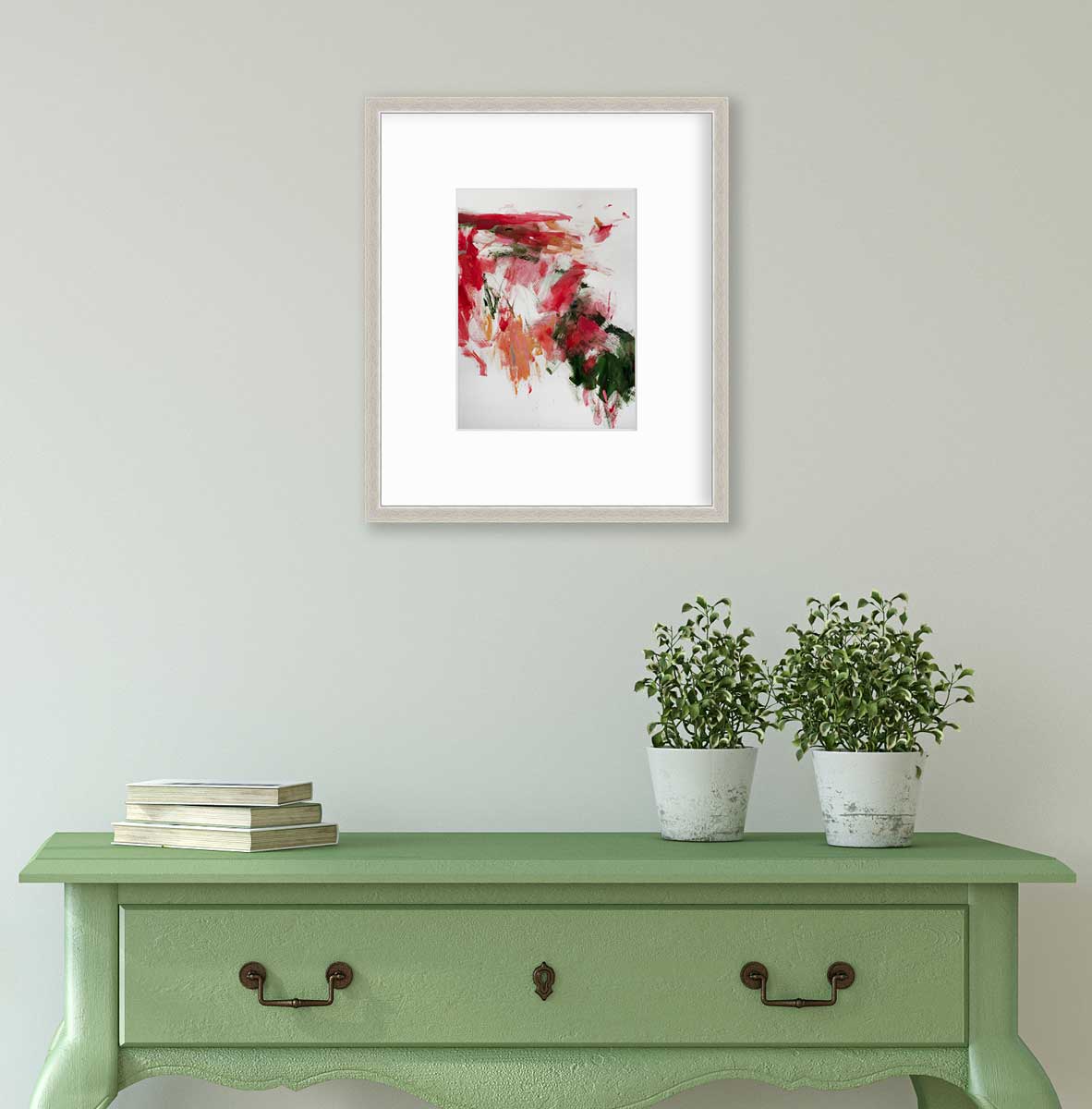 A red, green and white abstract print in a silver frame with a mat hangs on a white wall above a green console table with a stack of books and two potted plants.