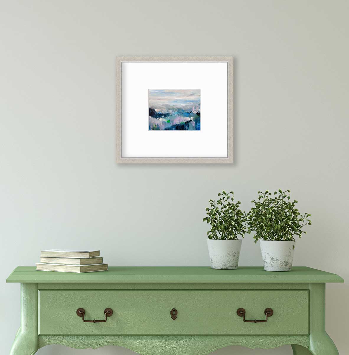 A blue abstract landscape by Kelly Rossetti in a silver frame with a mat hangs on a white wall above a green console table with a stack of books and two potted plants.