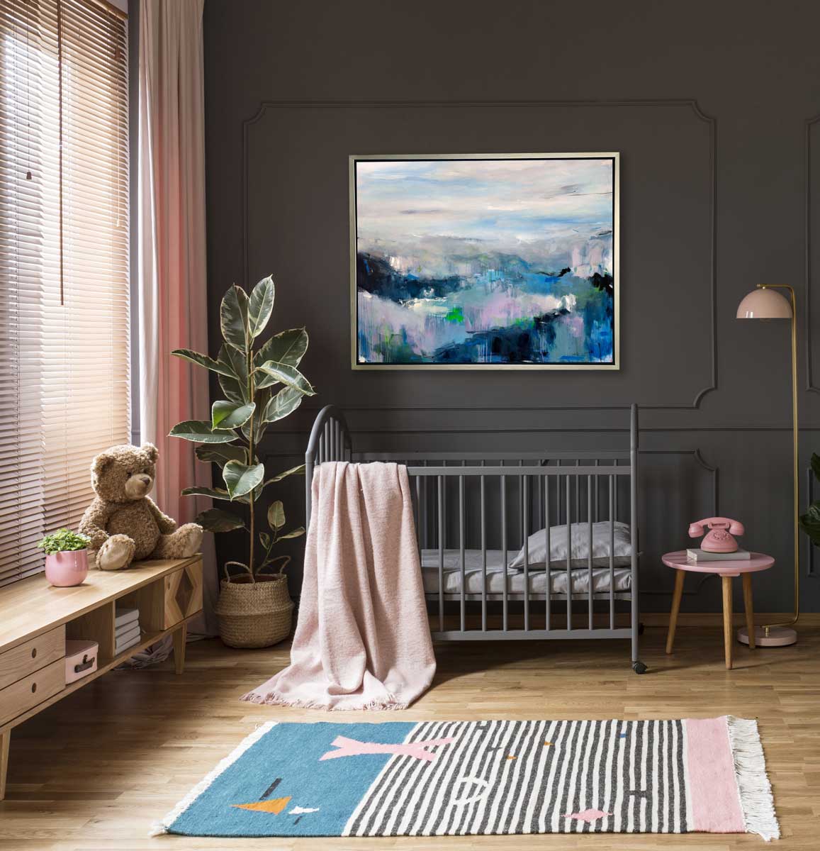 A blue abstract landscape print in a silver floater frame on a grey paneled wall above a grey crib with a blush colored blanket over the side, a low wooden cabinet with a teddy bear and a floor plant to the left and a small pink side table with a pink telephone to the right.