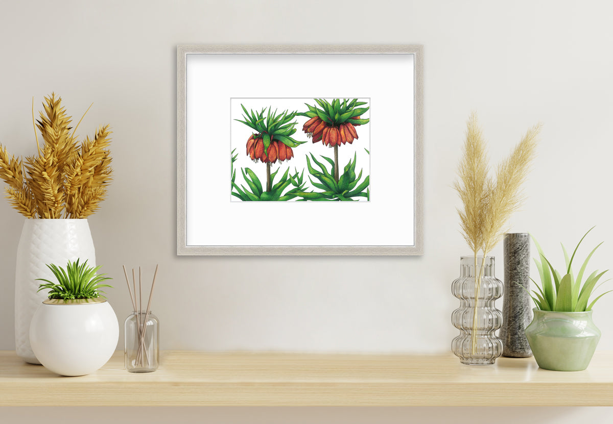 A green and red-orange floral print in a silver frame with a mat hangs on a white wall above a wood surface with several vases of potted plants.