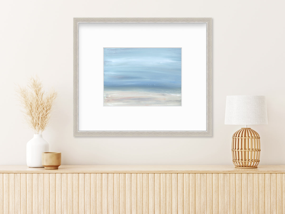 A white, blue and beige abstract seascape print in a silver frame with a mat hangs on a white wall above a natural wood surface with a wheat filled white vase, a tan decorative cup and a natural wood birdcage table lamp.