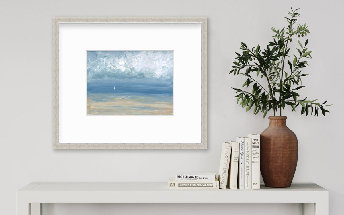 A white, blue and beige abstract seascape print in a silver frame with a mat hangs on a white wall above a white console table with a stack of books and a terracotta vase with greenery stems.