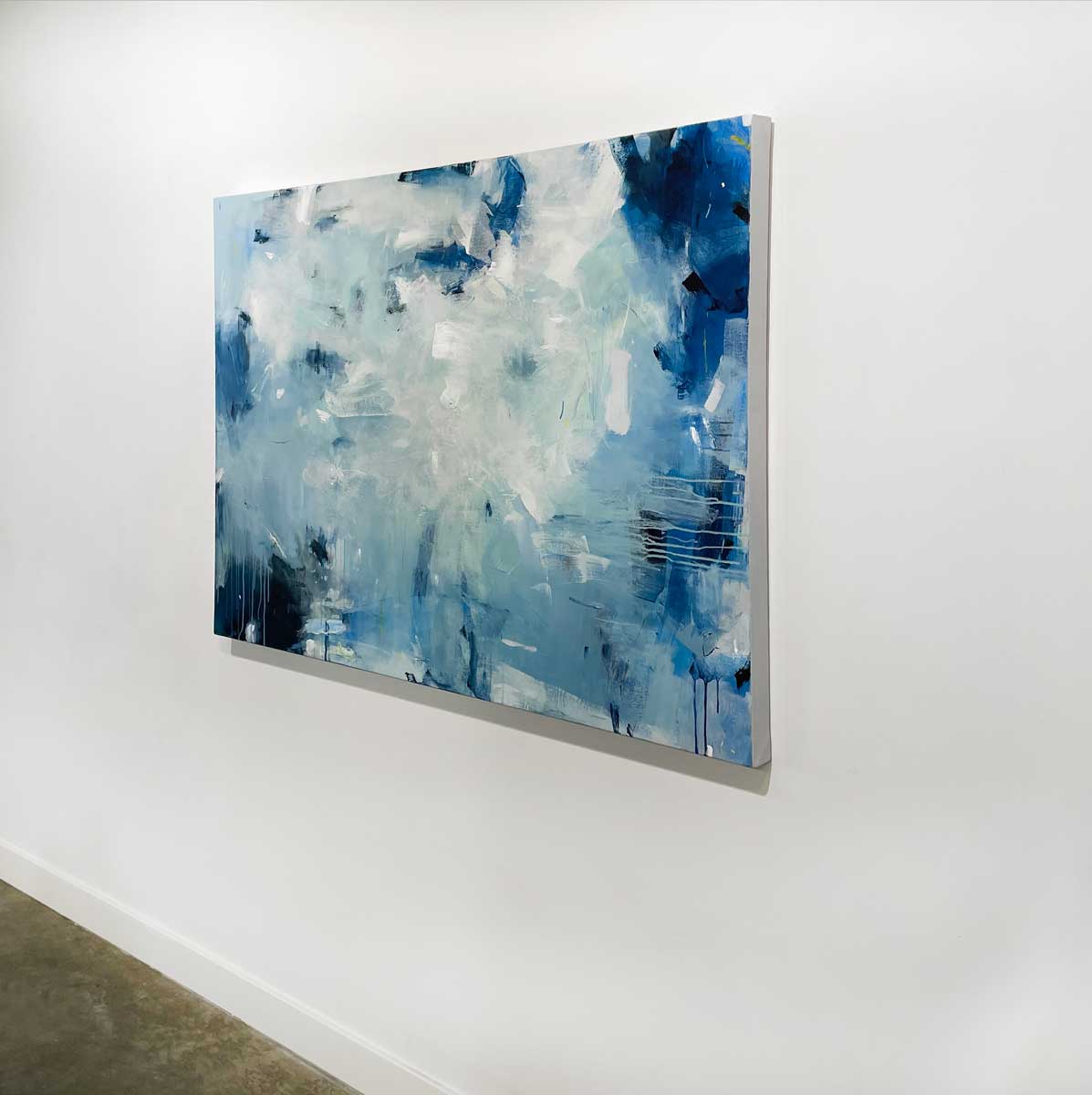 A right side-angle view of a blue and white abstract painting by Kelly Rossetti hangs on a white wall.
