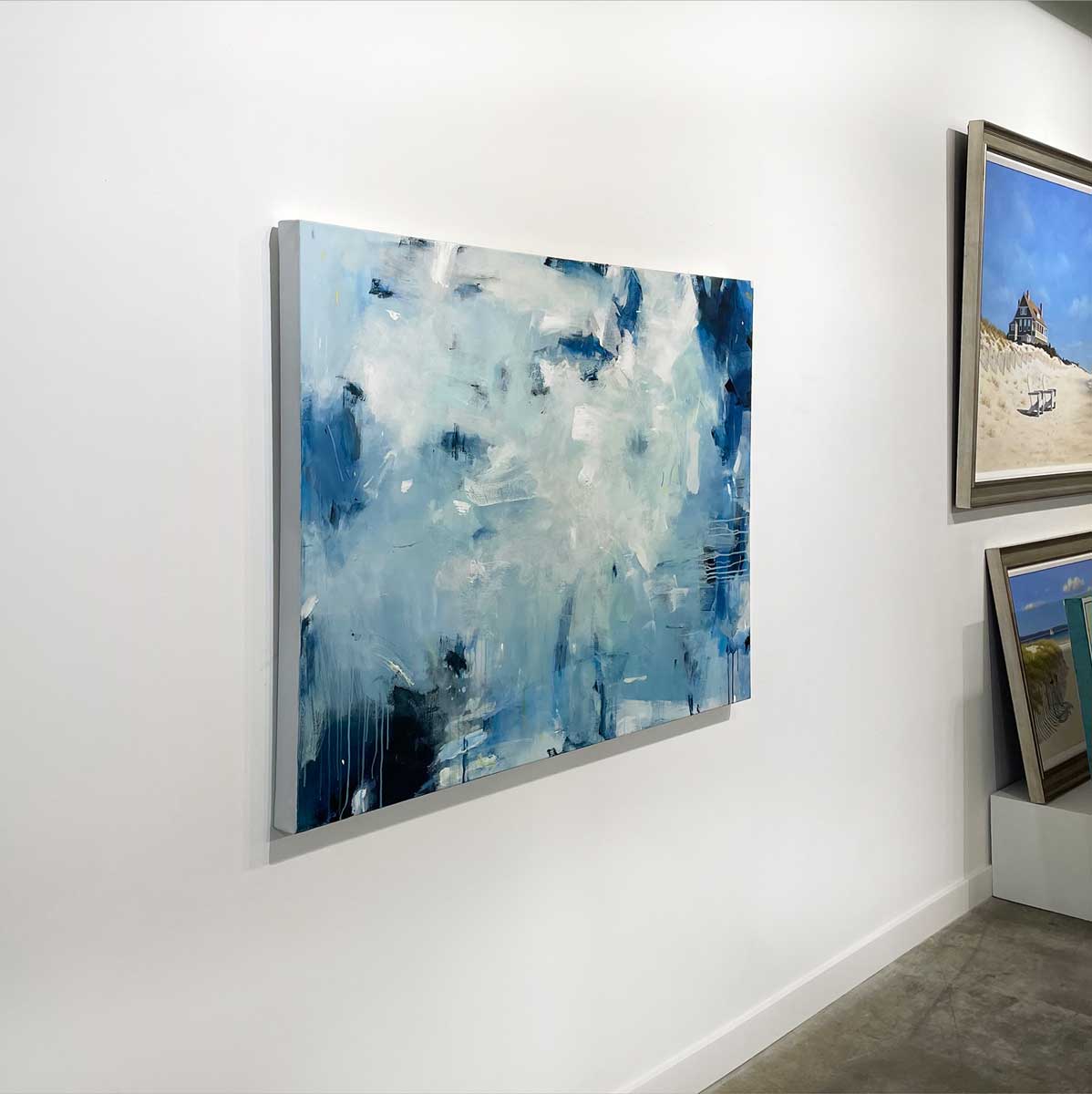A left side-angle view of a blue and white abstract painting hangs on a white wall in a gallery.