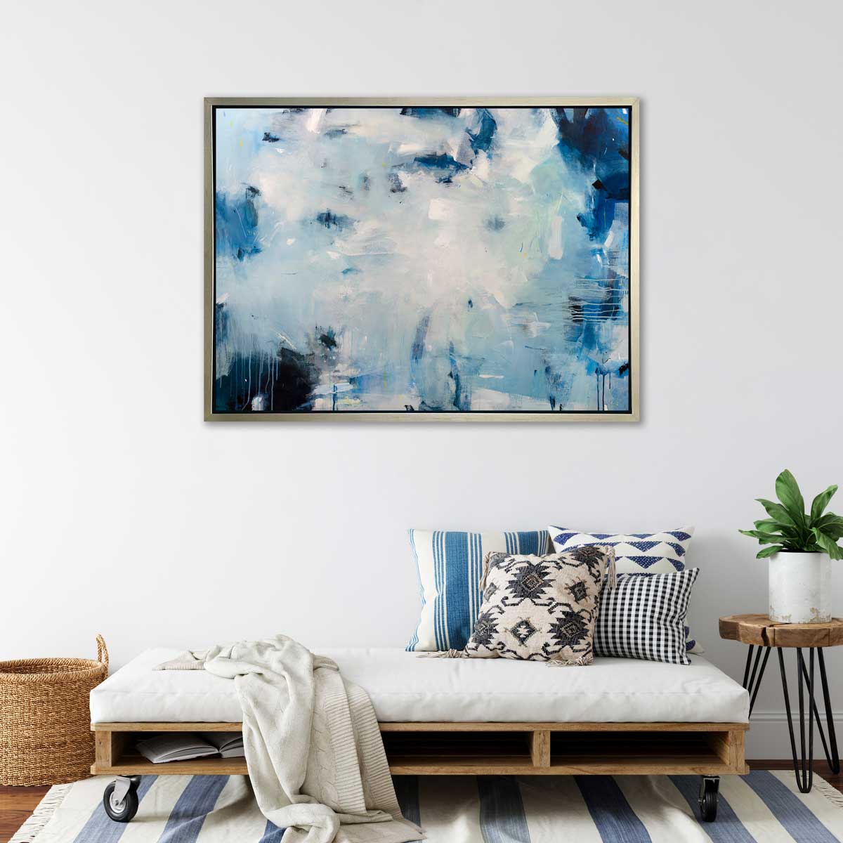 A blue, white and black abstract print in a silver floater frame hangs on a white wall above a wheeled bench with a white cushion and printed pillows, a live edge side table with a potted plant and a woven basket.