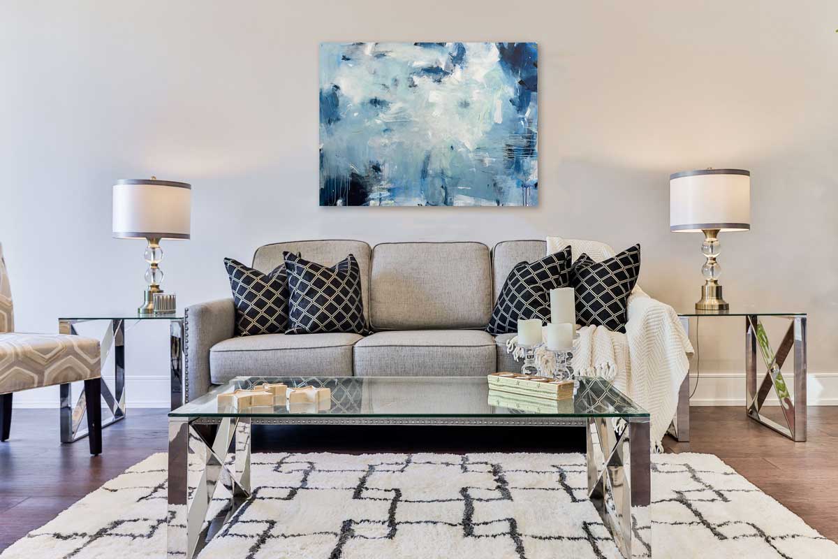 A blue abstract painting by Kelly Rossetti hangs on a white wall above a grey sofa with black geometric print pillows, two side tables with lamps and a rectangular glass top coffee table.